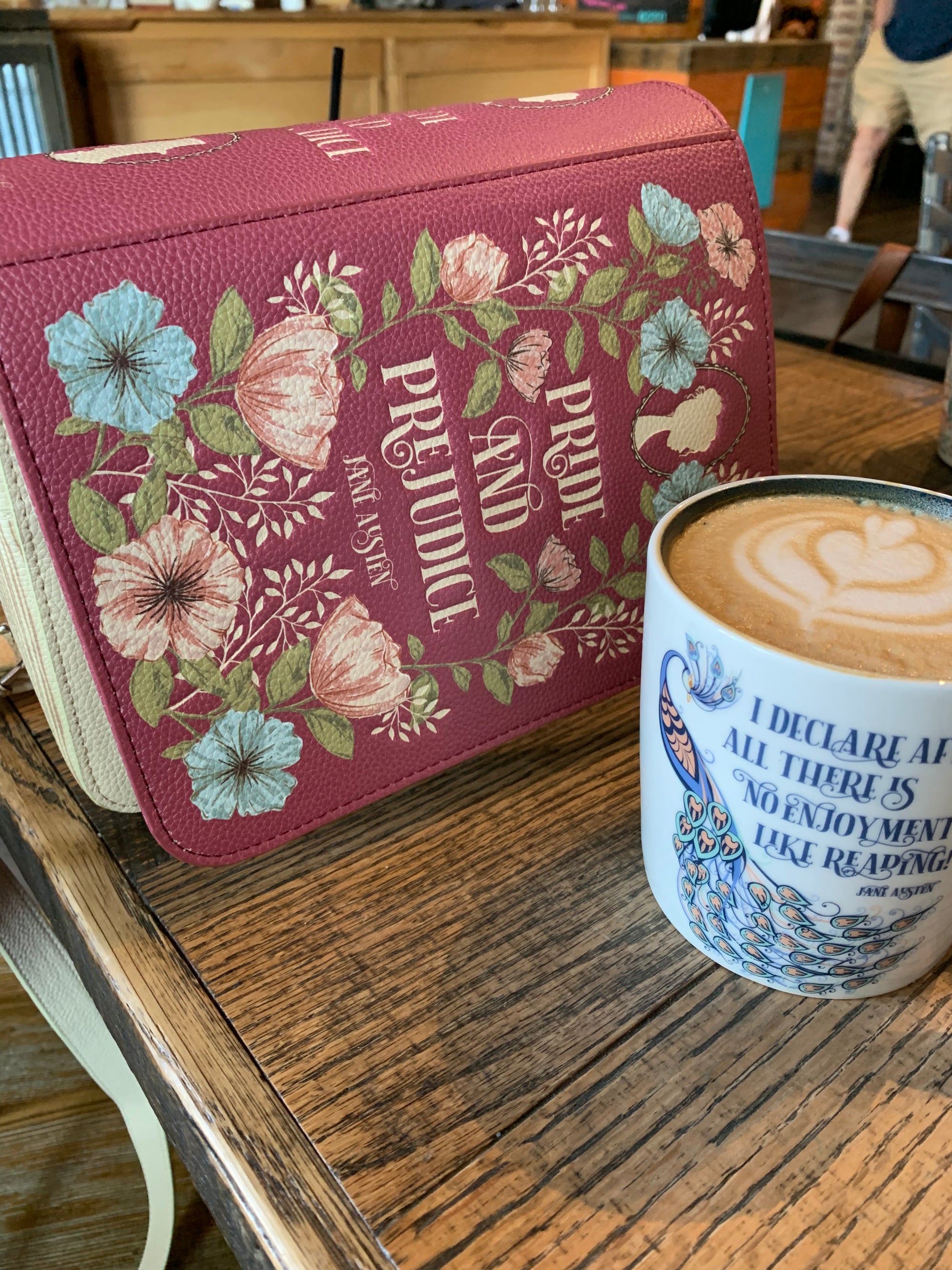 Crossbody with titled 'Pride and Prejudice' with floral design on cover, next to a cup of coffee with decorative sleeve.