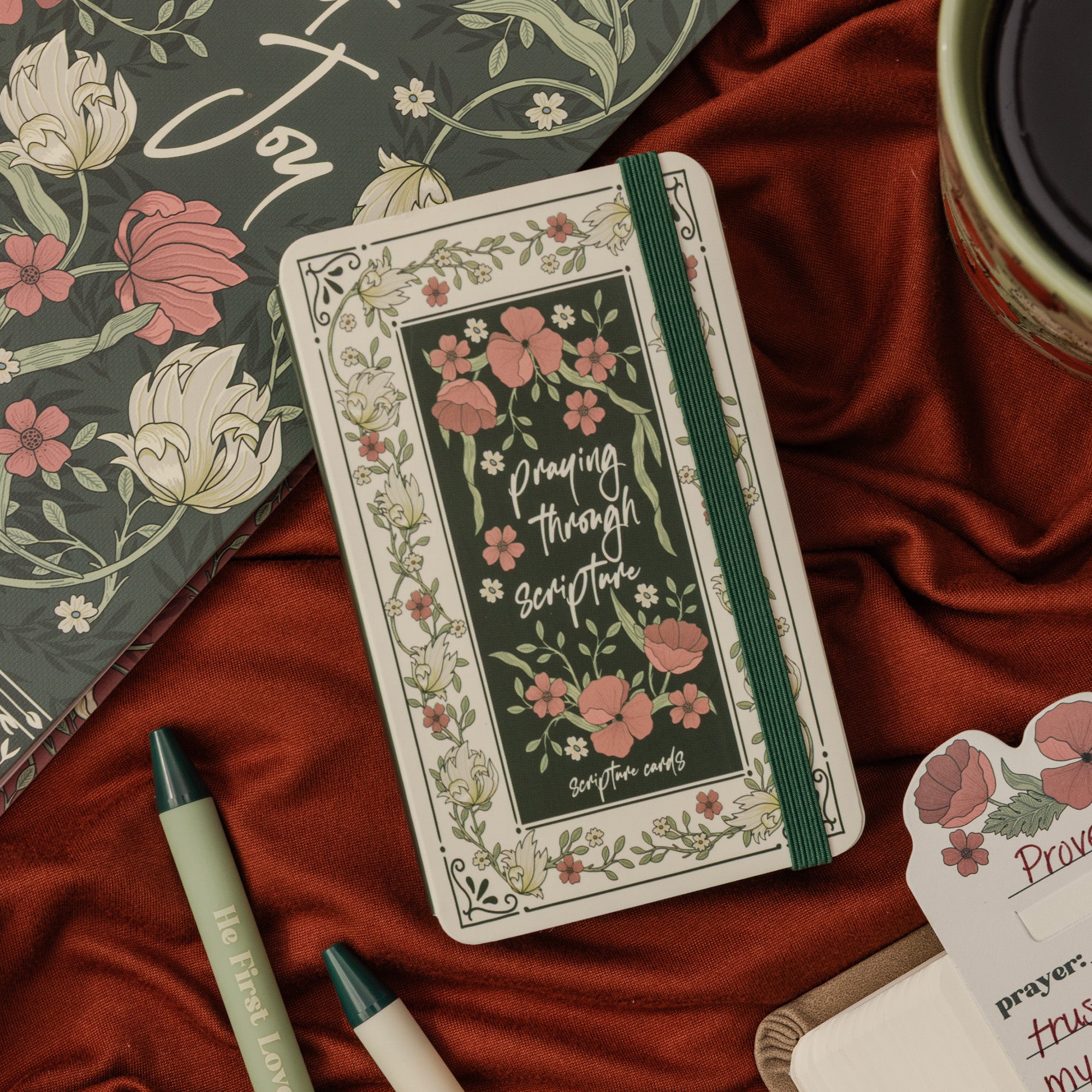 A rectangular prayer notebook in a white tin with a floral border and a dark green center panel reading 'Praying through scripture,' styled on red satin with a matching floral notebook, pens, and a cup nearby.