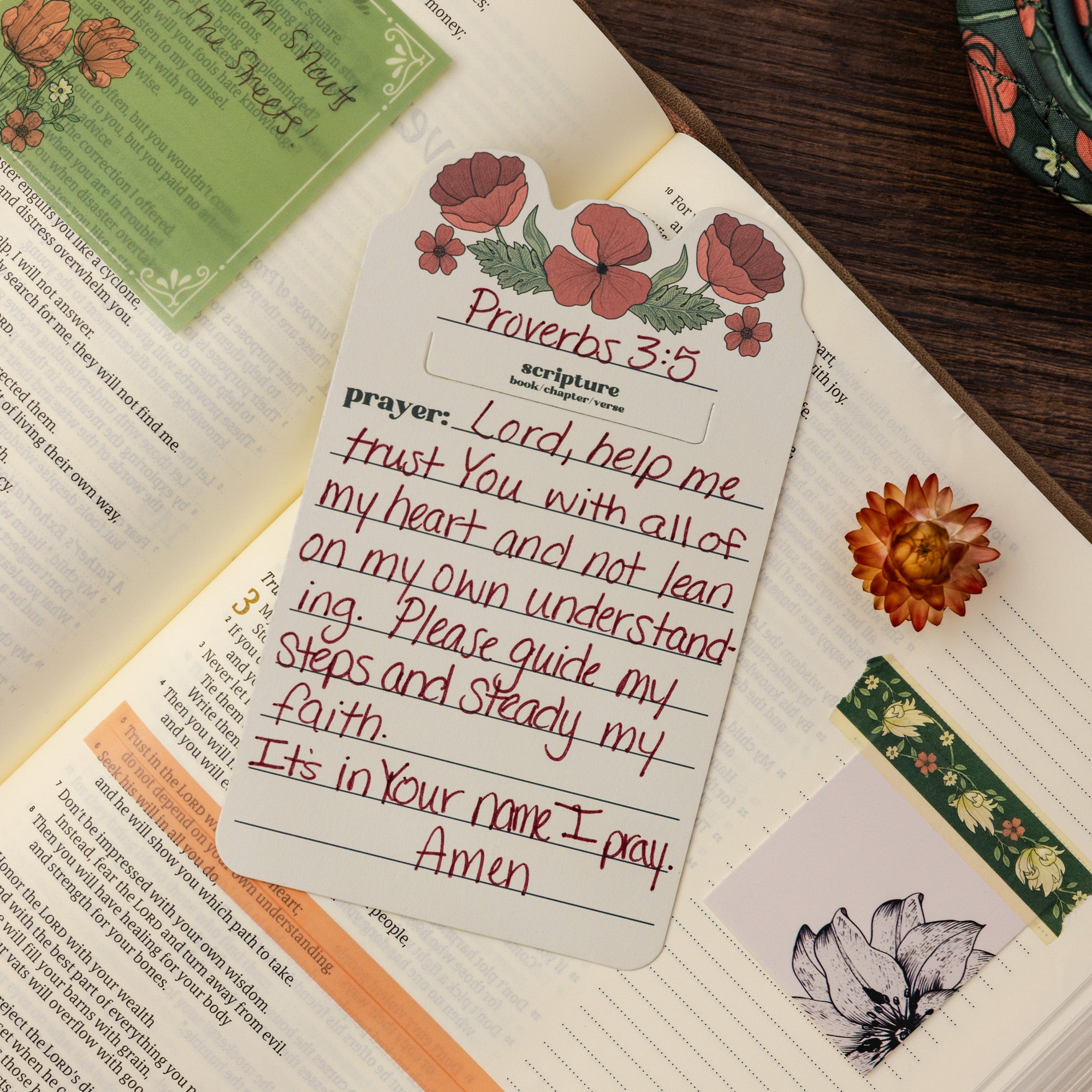 A floral bookmark or prayer card with Proverbs 3:5 written on it, styled on top of an open Bible with handwritten notes, a small dried flower, and decorative strips on a dark wood surface.