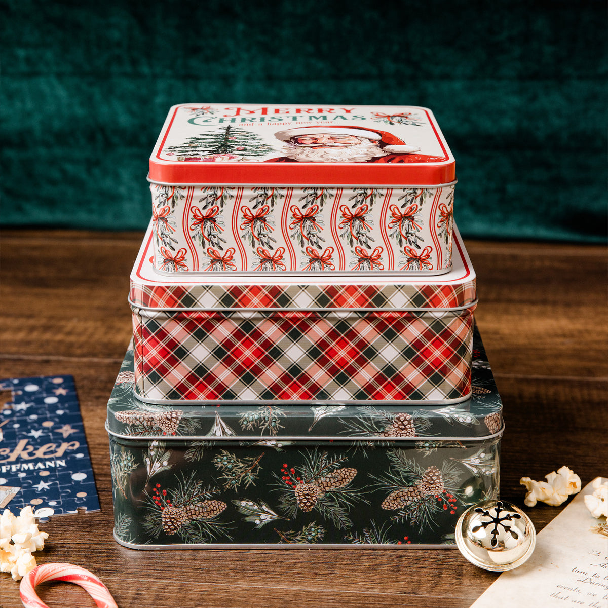 A stack of three decorative Christmas metal tins on a wooden surface, with a dark green velvet background. The top tin features a red and white Santa design with 'Merry Christmas' text, the middle tin has a red, black, and white plaid pattern, and the bottom tin is dark green with pine and berry motifs.