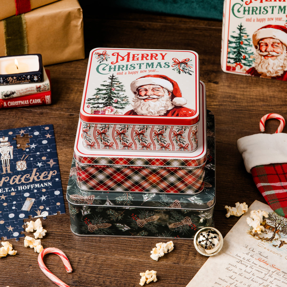 A stack of three decorative Christmas metal tins sits on a wooden tabletop surrounded by holiday props, including candy canes, popcorn, a blue nutcracker card, a candle, a plaid stocking, and another Santa-themed box in the background.