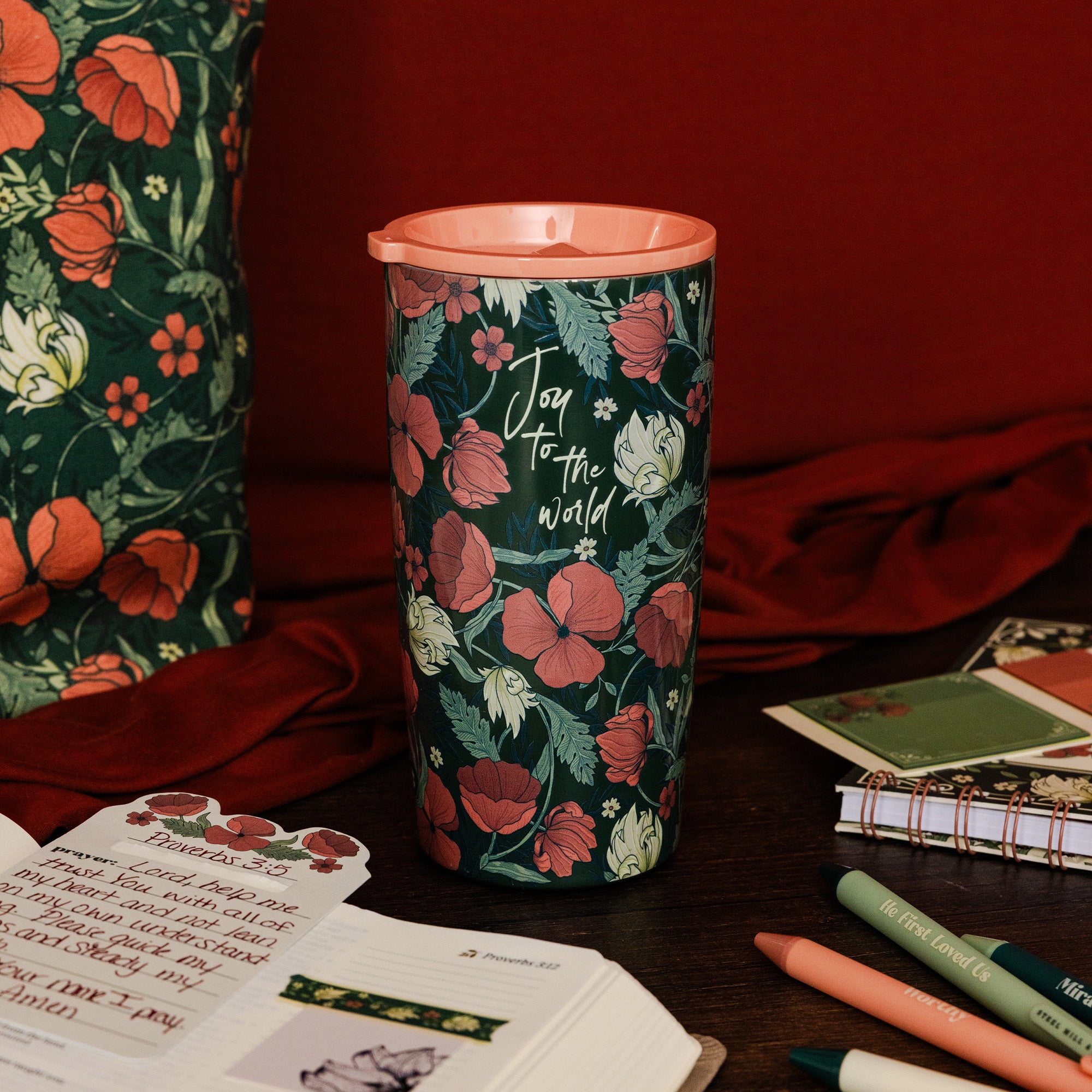 A tall insulated tumbler with a floral print and the words 'Joy to the world' sits on a dark wooden table surrounded by notebooks, pens, and a matching floral bag against a deep red fabric backdrop.