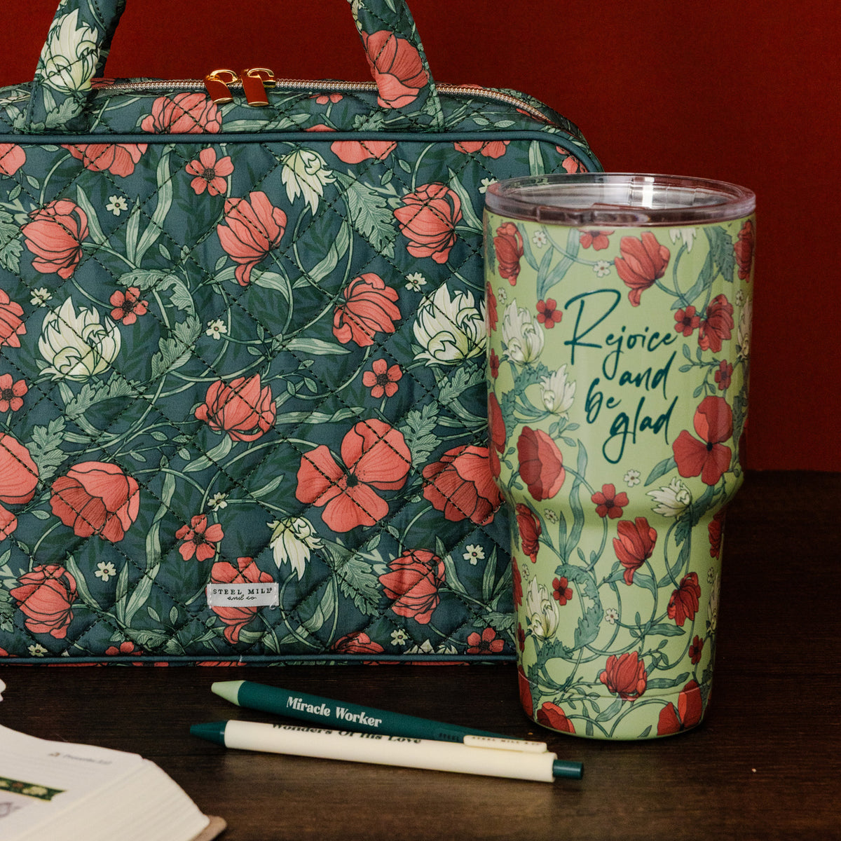 A quilted tote bag and a insulated tumbler with a matching floral print sit on a dark wooden surface against a dark red background. The tote is dark teal with coral and white floral patterns, and the tumbler is light green with the phrase 'Rejoice and be glad' in script.