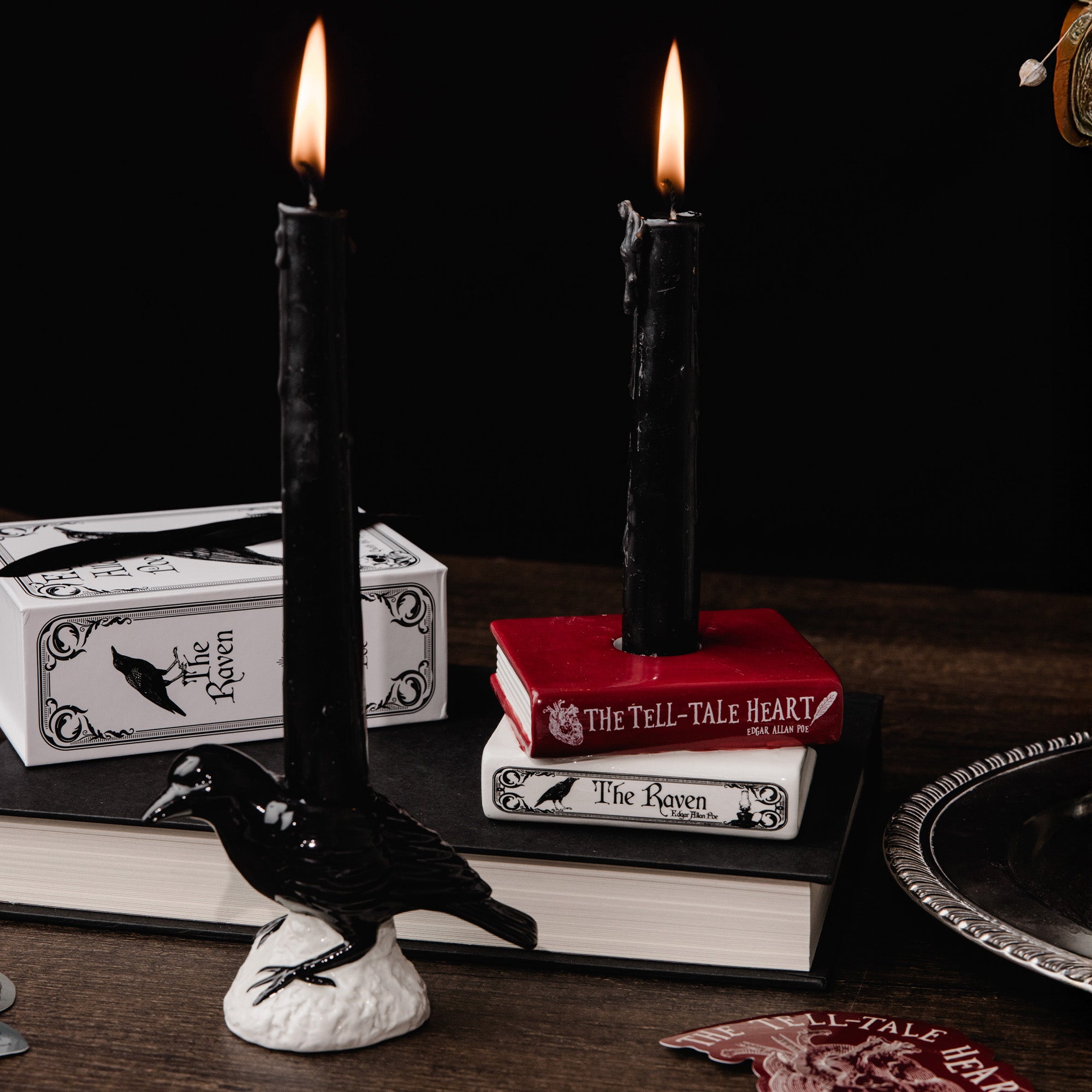 A gothic-themed tabletop scene featuring two black taper candles, one in a raven-shaped holder and one in a red book-shaped holder, alongside books and a silver tray on a dark wooden surface against a black background.