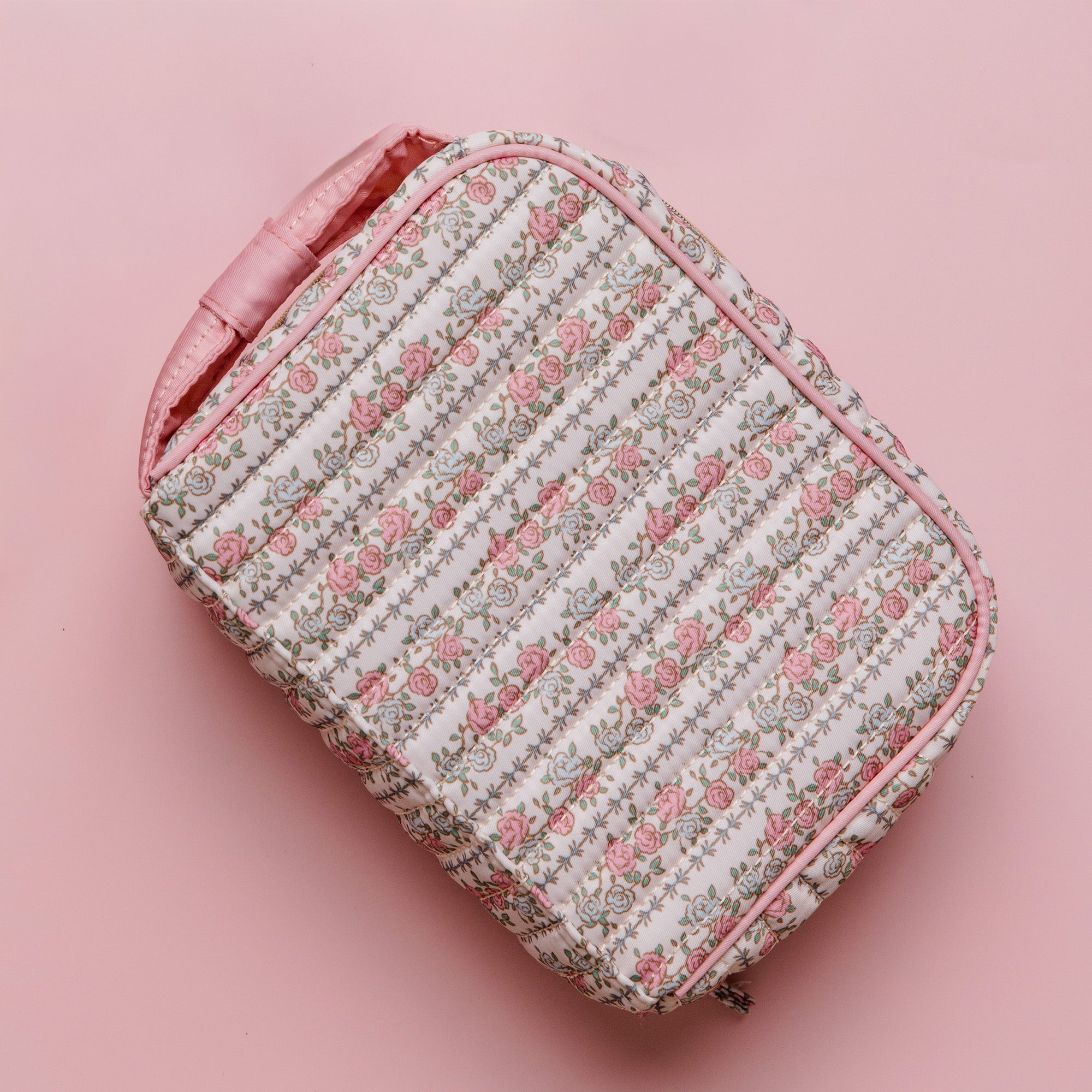 A quilted cosmetic bag with a pink floral pattern on a white background, featuring a pink handle and pink piping, set against a solid pink background.