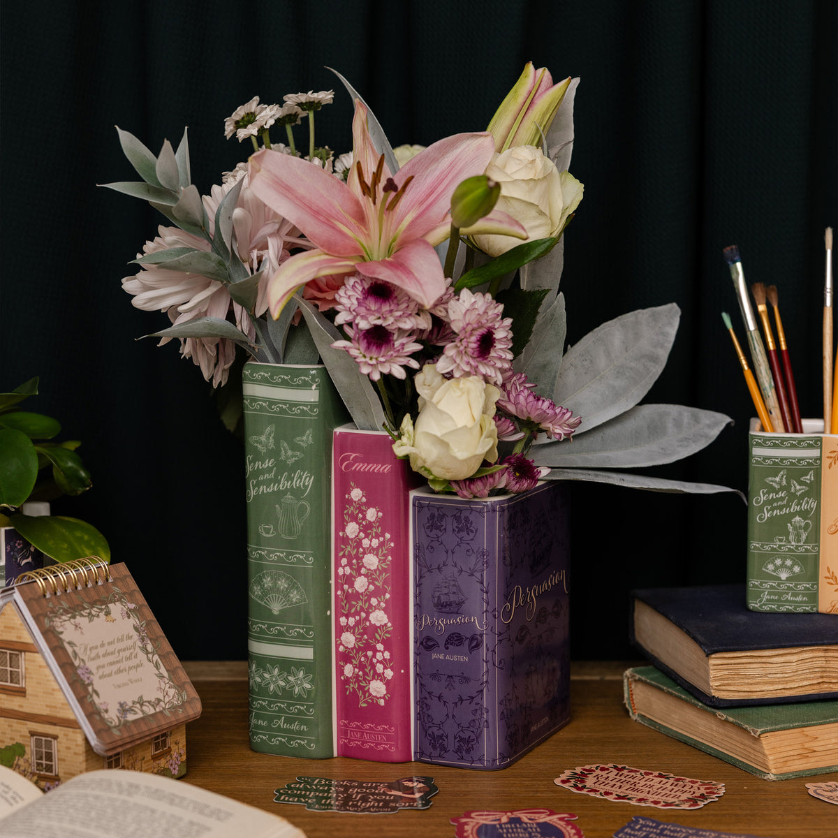 planter with three 'books' side by side. large green sense and sensibility, medium pink emma, and small purple persuasion on a table with a dark background
