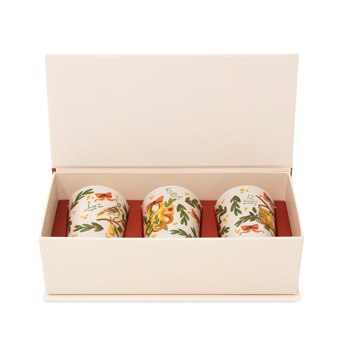 Three ceramic mugs with holiday illustrations displayed inside an open cream-colored gift box with a red interior.