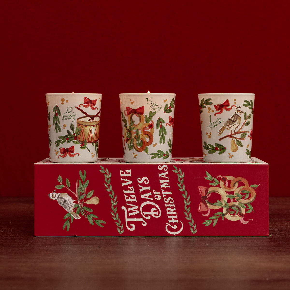 Three Christmas-themed candles displayed on top of a red gift box. The candles are cream-colored with festive illustrations including a drum, a snake, and a bird, decorated with red bows and green holly leaves.