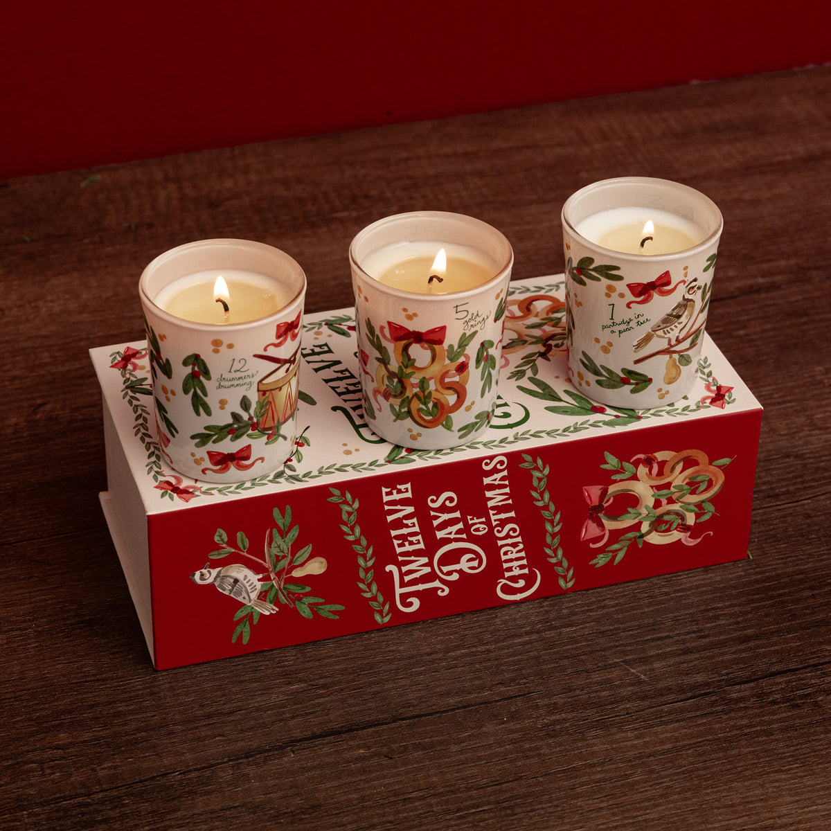 Three lit cream-colored candles in white glass holders with red and green Christmas illustrations, arranged on top of a red gift box printed with 'The Twelve Days of Christmas' text and holiday graphics, set on a dark wooden surface.