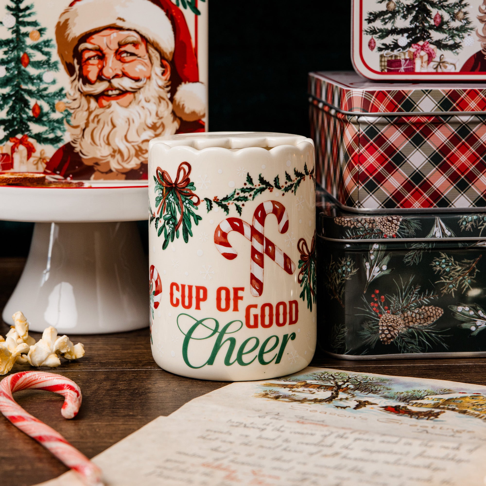 A ceramic Christmas mug with 'CUP OF GOOD Cheer' printed in red and green, decorated with candy canes and holly, set on a wooden table with holiday-themed props including candy canes, popcorn, and vintage-style tin boxes.