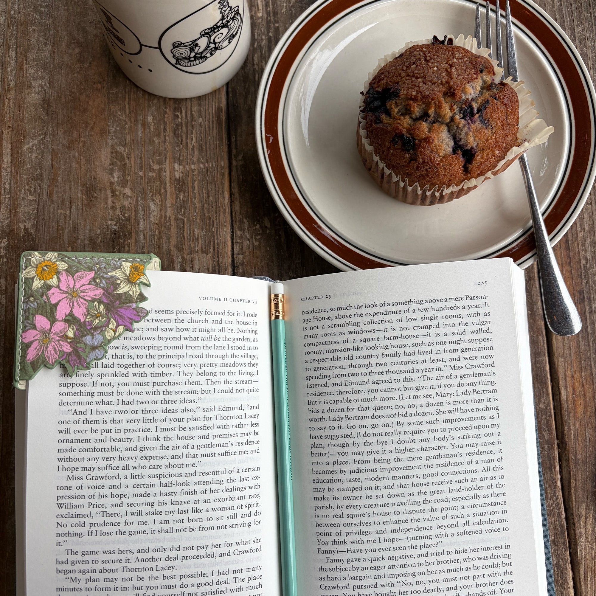 An open book spread with printed text and a pink floral bookmark on the left, alongside a plate with a brown pastry and a silver spoon on a wooden table.