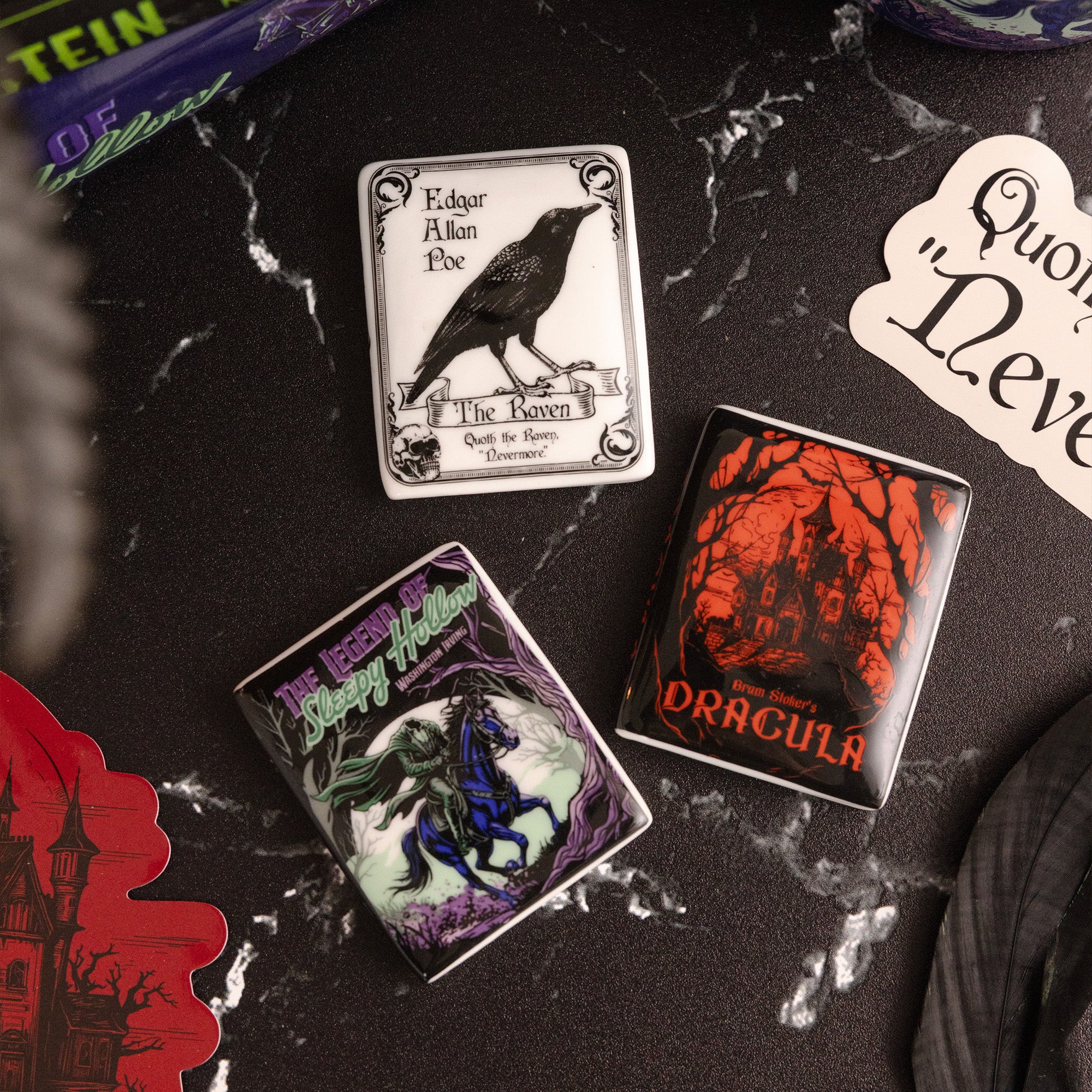 Three gothic-themed magnets with illustrated covers are arranged on a dark marble surface. The designs feature a raven on a white tin, a purple-and-green 'The Witch's Sleepy Hound' tin, and a red-and-black 'Dracula' tin.