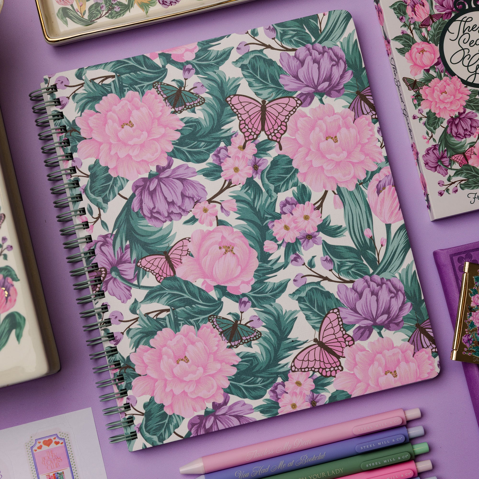 Floral notebook with pink flowers and butterflies on a purple surface with stationery items.