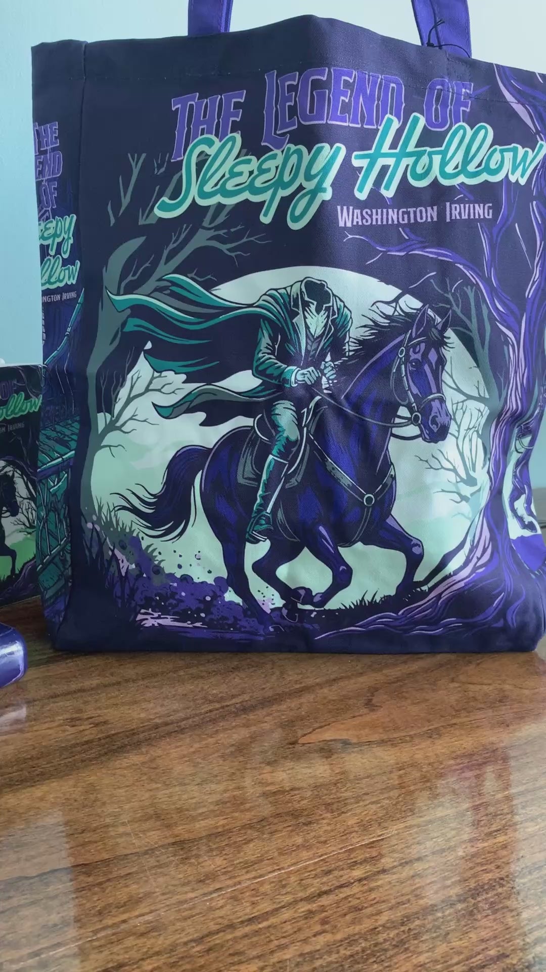 model showcasing dark tote bag with purple handles and a graphic print of a ghostly rider on a horse in a spooky forest, featuring the text 'The Legend of Sleepy Hollow' and 'Washington Irving'.