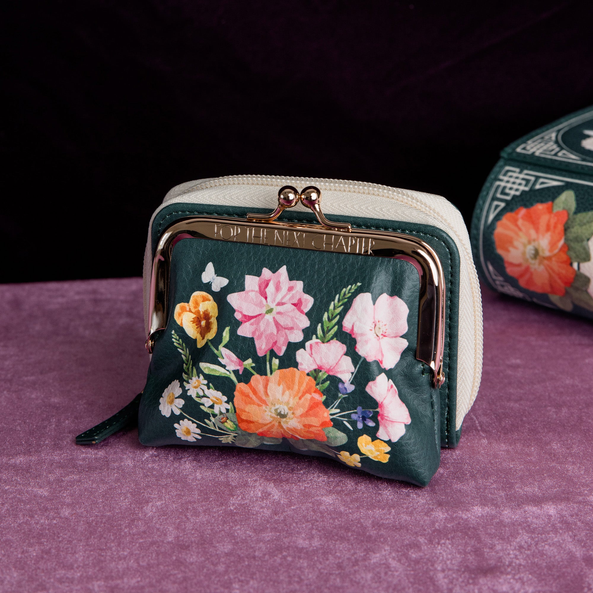 A small kiss-lock coin purse with a cream-colored frame and zipper, featuring a dark green front panel decorated with a colorful floral print of pink, orange, yellow, and white flowers on a purple base and black ground